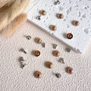 May include: An assortment of metal flower-shaped rivets and studs in gold and silver tones. The rivets have a fluted design, and the studs have a flat back. Some have a post for attachment. The items are scattered on a textured white surface.