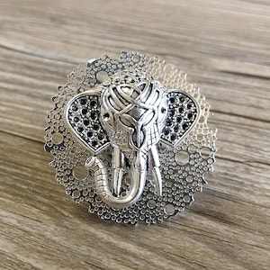 以下が含まれることがあります： 複雑なディテールが施された銀色の金属製の象のチャームが、円形のフィリグリーデザインの中にセットされています。象の頭は前を向いており、鼻は持ち上げられています。