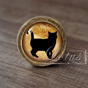 Może przedstawiać: Okrągła gałka do szafki z czarną sylwetką kota na złotym i brązowym tle. Gałka ma brązową metalową obwódkę i jest osadzona na drewnianej powierzchni. Gałka ma średnicę około 3,8 cm.
