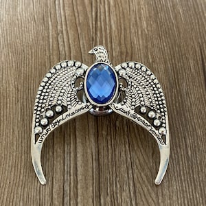 Peut inclure: Un oiseau argenté aux ailes déployées, tenant une pierre précieuse bleue dans son bec. Les ailes sont ornées de petites perles et l'inscription "What's Beyond Measure" est visible sur l'aile gauche.