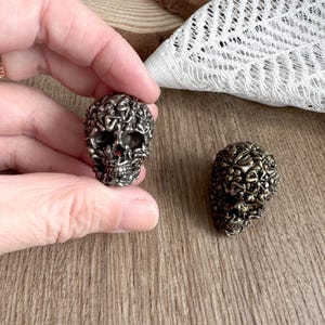 May include: Two metal skull figurines. One is held in a hand, the other rests on a wooden surface. The skulls have a detailed, textured design, with a dark, metallic finish. The skulls are a unique decorative item.