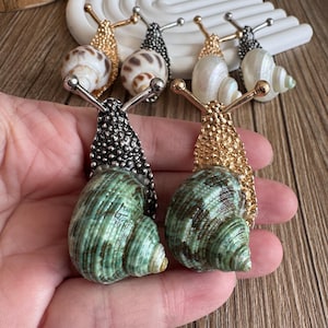 以下が含まれることがあります： 金属のアクセントが付いたカタツムリ型のブローチのコレクション。ブローチは、白、茶色、緑など、さまざまな貝殻のデザインが特徴で、金、銀、黒の金属のディテールが施されています。カタツムリの触角も金属製です。