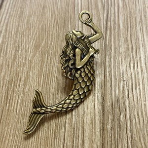 K&ouml;nnte beinhalten: Ein goldfarbener Meerjungfrauen-Anh&auml;nger mit detailliertem Design. Die Meerjungfrau h&auml;lt einen Ring in der Hand und hat lange, flie&szlig;ende Haare. Der Anh&auml;nger ist perfekt, um Schmuck oder Bastelarbeiten eine Prise Verspieltheit zu verleihen.