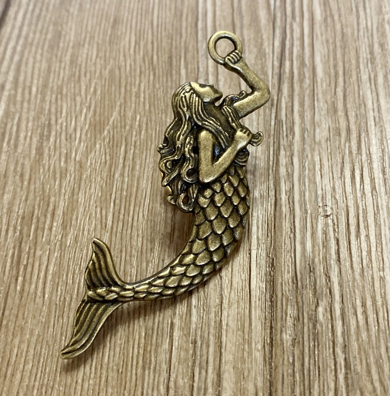 K&ouml;nnte beinhalten: Ein goldfarbener Meerjungfrauen-Anh&auml;nger mit detailliertem Design. Die Meerjungfrau h&auml;lt einen Ring in der Hand und hat lange, flie&szlig;ende Haare. Der Anh&auml;nger ist perfekt, um Schmuck oder Bastelarbeiten eine Prise Verspieltheit zu verleihen.