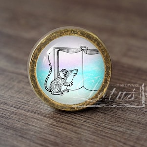 Può includere: Un pomello rotondo in ottone con un inserto in vetro che presenta un'illustrazione in bianco e nero di un topo che guarda un libro aperto su uno sfondo blu e rosa sfumato.