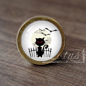 Pode incluir: Um gato preto com olhos vermelhos senta-se em uma cerca na frente de uma lua cheia. Os morcegos voam por cima. A imagem está em um botão redondo de ouro antigo.