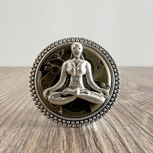 Peut inclure: Une bague en argent avec un motif détaillé d'une personne en position de méditation assise sur un fond circulaire avec des engrenages et des rouages.