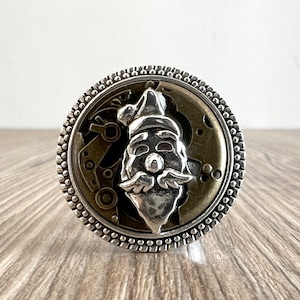 May include: A silver-toned ring featuring a Santa Claus face design. The ring's face has a detailed, textured Santa Claus figure set against a backdrop of watch gears. The ring has a beaded border.