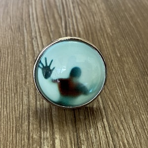 Puede incluir: Un anillo de plata con un cabujón de vidrio transparente que presenta la silueta de una persona con la mano extendida. La persona es de color azul oscuro y marrón.