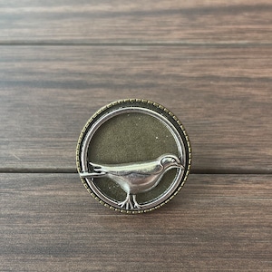 Op de afbeelding: Een zilveren vogel in een gouden rond frame. De vogel bevindt zich in een vliegende houding met uitgespreide vleugels. Het frame heeft een getextureerde rand.