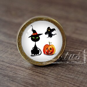 Pode incluir: Um botão de armário redondo de cor dourada antiga com um fundo branco e um design temático de Halloween. O design apresenta um gato preto com um chapéu de bruxa, um fantasma e uma abóbora.