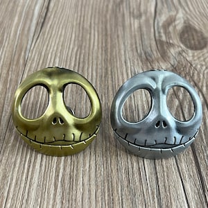 Könnte beinhalten: Zwei Jack-Skellington-Schubladenknöpfe aus Metall, einer in Gold und einer in Silber. Die Knöpfe sind in Form von Jack Skellingtons Gesicht mit einem genähten Lächeln gestaltet.