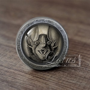 May include: A silver knob with a round, glass face featuring a black and white illustration of a bat with its mouth open.