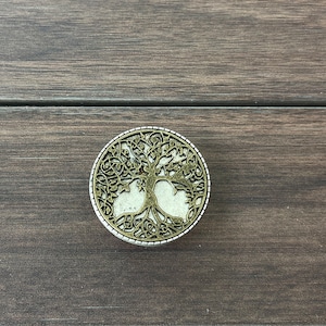 May include: A round, silver-colored metal ornament with a tree of life design. The tree is cut out of the metal, revealing a white background.