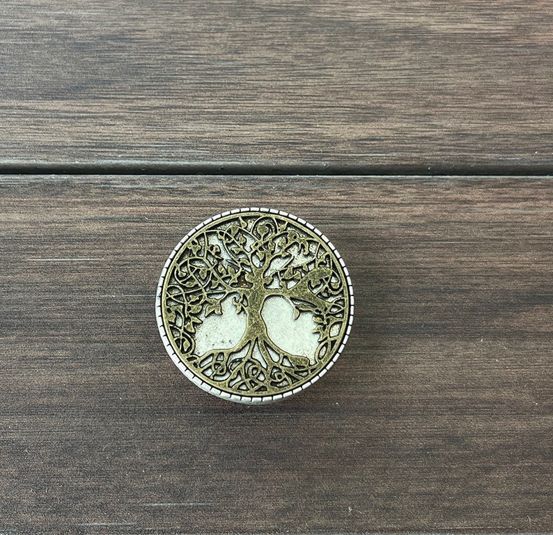 May include: A round, silver-colored metal ornament with a tree of life design. The tree is cut out of the metal, revealing a white background.