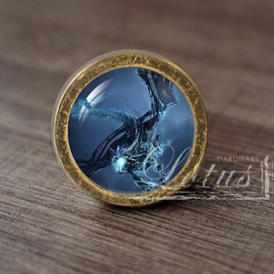 May include: A round, brass-colored cabinet knob with a glass insert featuring a blue dragon with glowing eyes against a dark blue and gray cloudy sky.