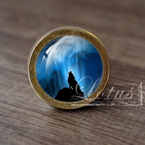 Può includere: Un pomello per armadio rotondo color ottone con un inserto in vetro che presenta una silhouette di un lupo che ulula alla luna. La luna è rappresentata come un globo blu e bianco brillante.