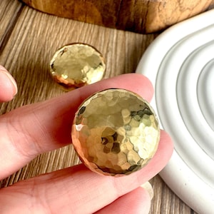 May include: Two hammered gold cabinet knobs with a textured surface. The knobs are round and have a shiny, metallic finish. The knobs are likely used for furniture or decorative purposes.