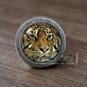 Może przedstawiać: Srebrny gałka do szafki z okrągłym, szklanym frontem przedstawiającym zbliżenie twarzy jaguara. Futro jaguara jest połączeniem brązu, czerni i złota.