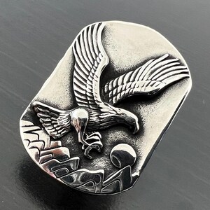 May include: A silver-toned rectangular belt buckle featuring a detailed relief of an eagle in flight. The eagle is depicted with outstretched wings above a mountain range and a rising sun. The buckle has a brushed metal finish.