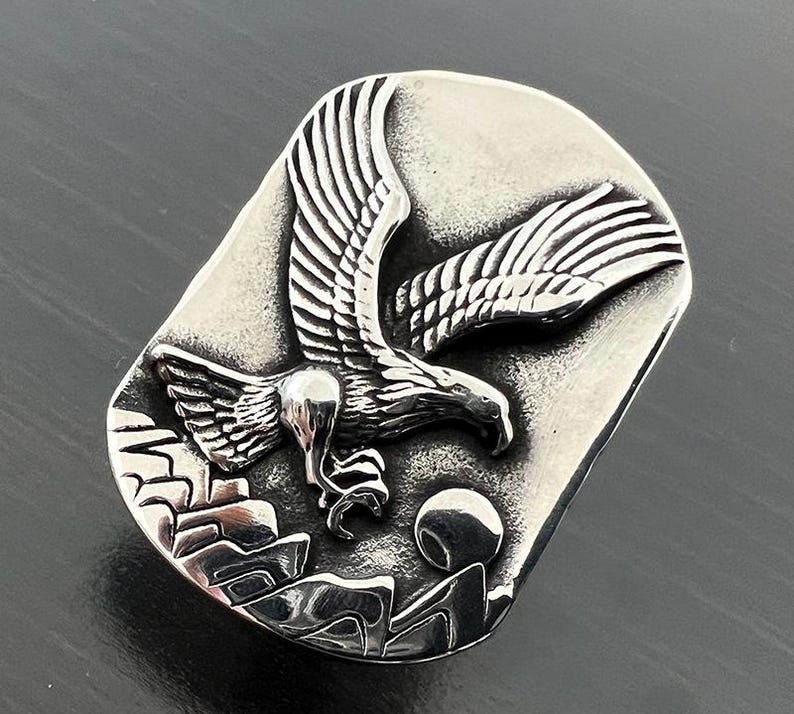 May include: A silver-toned rectangular belt buckle featuring a detailed relief of an eagle in flight. The eagle is depicted with outstretched wings above a mountain range and a rising sun. The buckle has a brushed metal finish.
