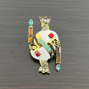 Può includere: Un fermaglio dorato con un designo in smalto bianco di un re che tiene uno scettro. Il fermaglio è decorato con strass rossi, blu e verdi.