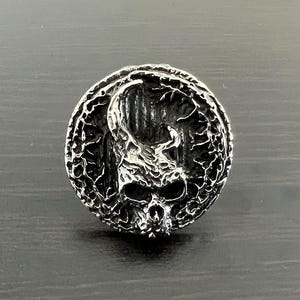 Puede incluir: Un anillo de plata con un diseño de calavera detallado. La calavera está rodeada por un marco circular texturizado.