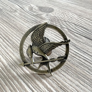 May include: A bronze-colored Mockingjay pin with a stylized arrow piercing through the bird's body. The pin is circular and features a detailed design of the Mockingjay with its wings spread.