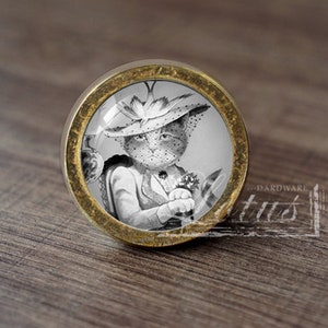 May include: A round, gold-toned cabinet knob with a glass insert featuring a black and white illustration of a cat wearing a hat and holding a flower.
