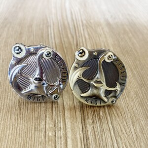 May include: A pair of metal cufflinks, one silver and one bronze. Each cufflink has a distinctive design featuring a stylized face, the text "Kiseiju" and "Migi". The cufflinks are circular with a detailed, mechanical appearance.