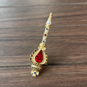 May include: A gold-toned brooch with a red teardrop-shaped stone and a long, slender stem adorned with white and blue beads.