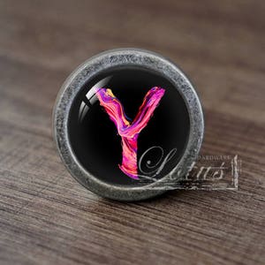 May include: A silver cabinet knob with a black background and a colourful, abstract letter Y painted on it.