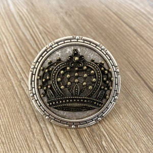 May include: A silver and gold decorative knob with a crown design. The knob has a textured surface and a raised crown with a beaded edge.