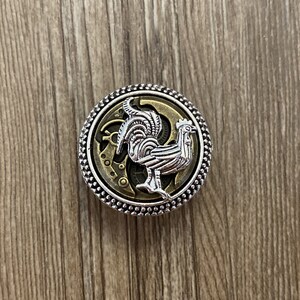 May include: A silver and gold metal button with a detailed design of a rooster with a clockwork background.