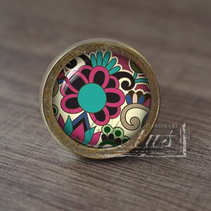 May include: A round, brass-coloured cabinet knob with a colourful floral design. The design features a large pink flower with a turquoise centre, surrounded by smaller flowers and swirls in shades of pink, blue, green, and black.