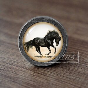 Op de afbeelding: Een ronde, zilverkleurige kastknop met een zwart paard dat op een beige achtergrond rent. De knop heeft een verhoogde, getextureerde rand.