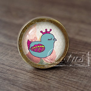 May include: A round, brass-colored cabinet knob with a glass insert featuring a cartoon blue bird wearing a pink crown. The bird is surrounded by pink and white flowers on a white background with a grid pattern.