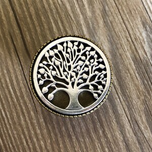 May include: A silver tree of life design with a dark brown background, surrounded by a gold-colored border.