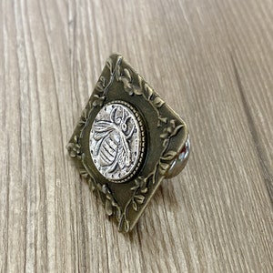 May include: A square-shaped, ornate, brass-colored drawer pull with a silver-colored bee design in the center.