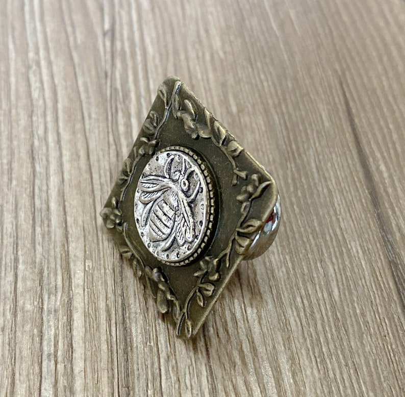 May include: A square-shaped, ornate, brass-colored drawer pull with a silver-colored bee design in the center.