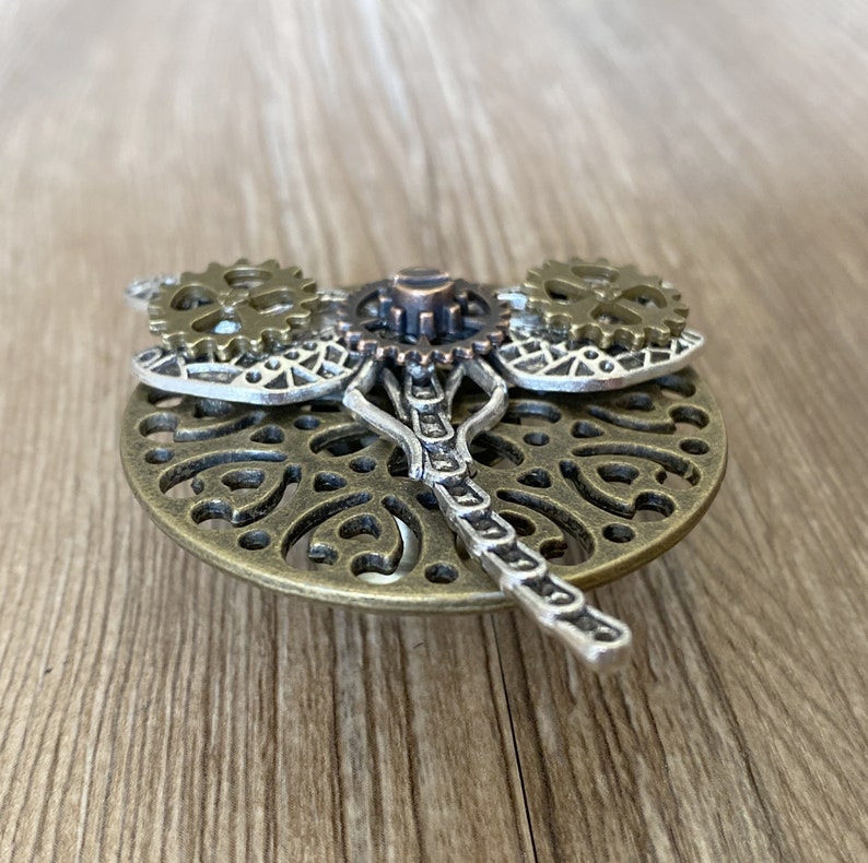 Może przedstawiać: Bransoletka w stylu steampunk w kształcie ważki, wykonana z brązu i srebra, z misternymi detalami i kołami zębatymi. Ważka ma filigranowy wz&oacute;r i łańcuszkowe ciało.