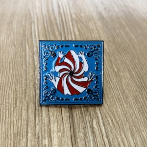 May include: A blue enamel pin with a white and red swirl design inside a square frame. The swirl design resembles a frog.
