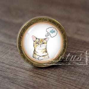 Może przedstawiać: Okrągła gałka do szafki z akwarelową ilustracją kota marzącego o rybie. Gałka ma podstawę z metalu w kolorze brązu i przezroczysty szklany lub żywiczny wierzch. Kot jest brązowo-beżowy z ponurym wyrazem twarzy.