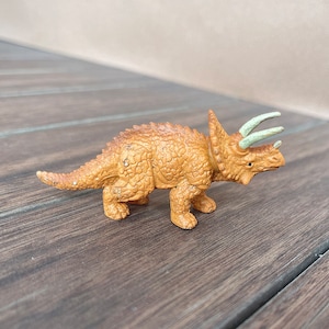 May include: A small, tan Triceratops figurine with a textured body, three horns, and a frill. The horns are a light green colour. The dinosaur toy is standing on a wooden surface.