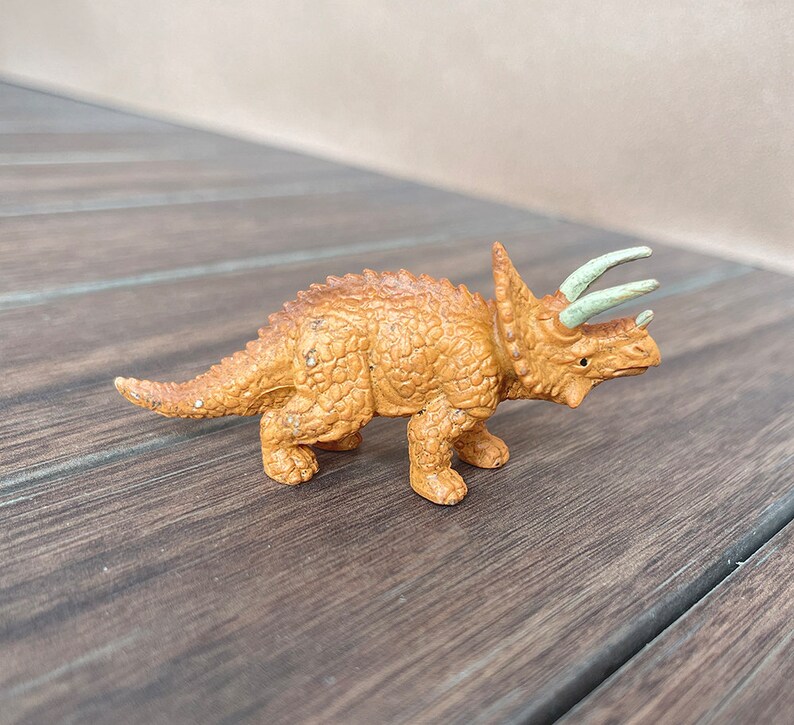 May include: A small, tan Triceratops figurine with a textured body, three horns, and a frill. The horns are a light green colour. The dinosaur toy is standing on a wooden surface.