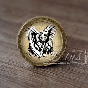 Op de afbeelding: Een ronde, messingkleurige kastknop met een glazen inzetstuk met een zwart-wit afbeelding van de Grim Reaper met een zeis en vleugels.