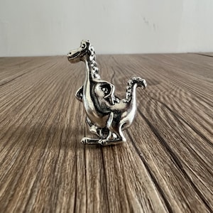 Pode incluir: Uma figurinha de dinossauro de cor prateada com uma superfície texturizada. O dinossauro está em pé sobre uma superfície de madeira.