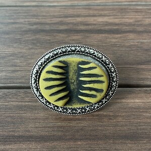 Può includere: Un anello ovale in argento con un design a motivi neri e gialli. L'anello ha un bordo decorativo con piccoli punti e croci argentati.