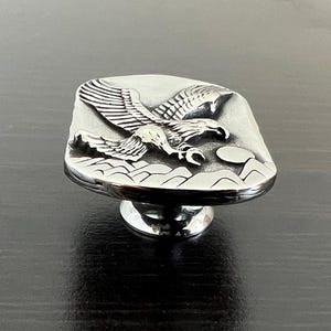 May include: A silver-toned cabinet knob with a detailed relief of an eagle in flight over a mountain range. The knob has a rectangular face and a round base, suitable for furniture or decor.