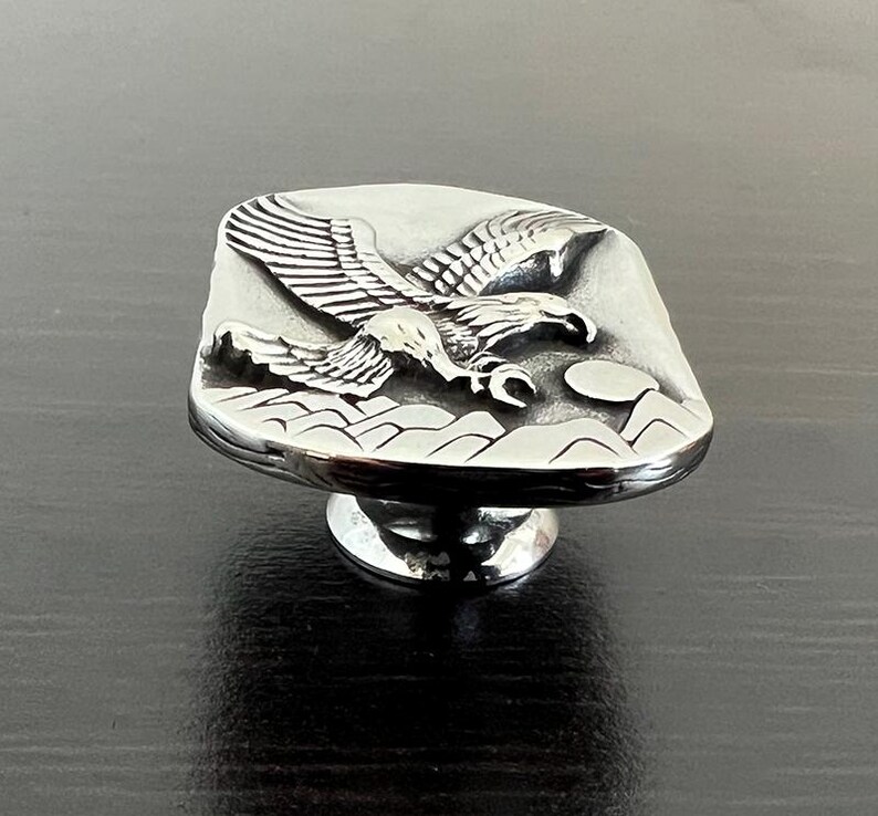 May include: A silver-toned cabinet knob with a detailed relief of an eagle in flight over a mountain range. The knob has a rectangular face and a round base, suitable for furniture or decor.
